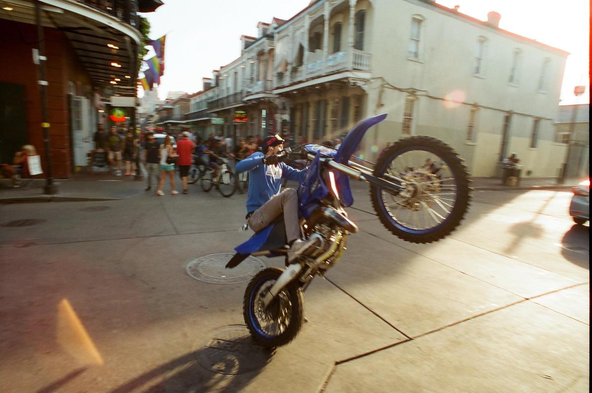 A man on a dirt bike performs a wheelie on a street lined with buildings and people.