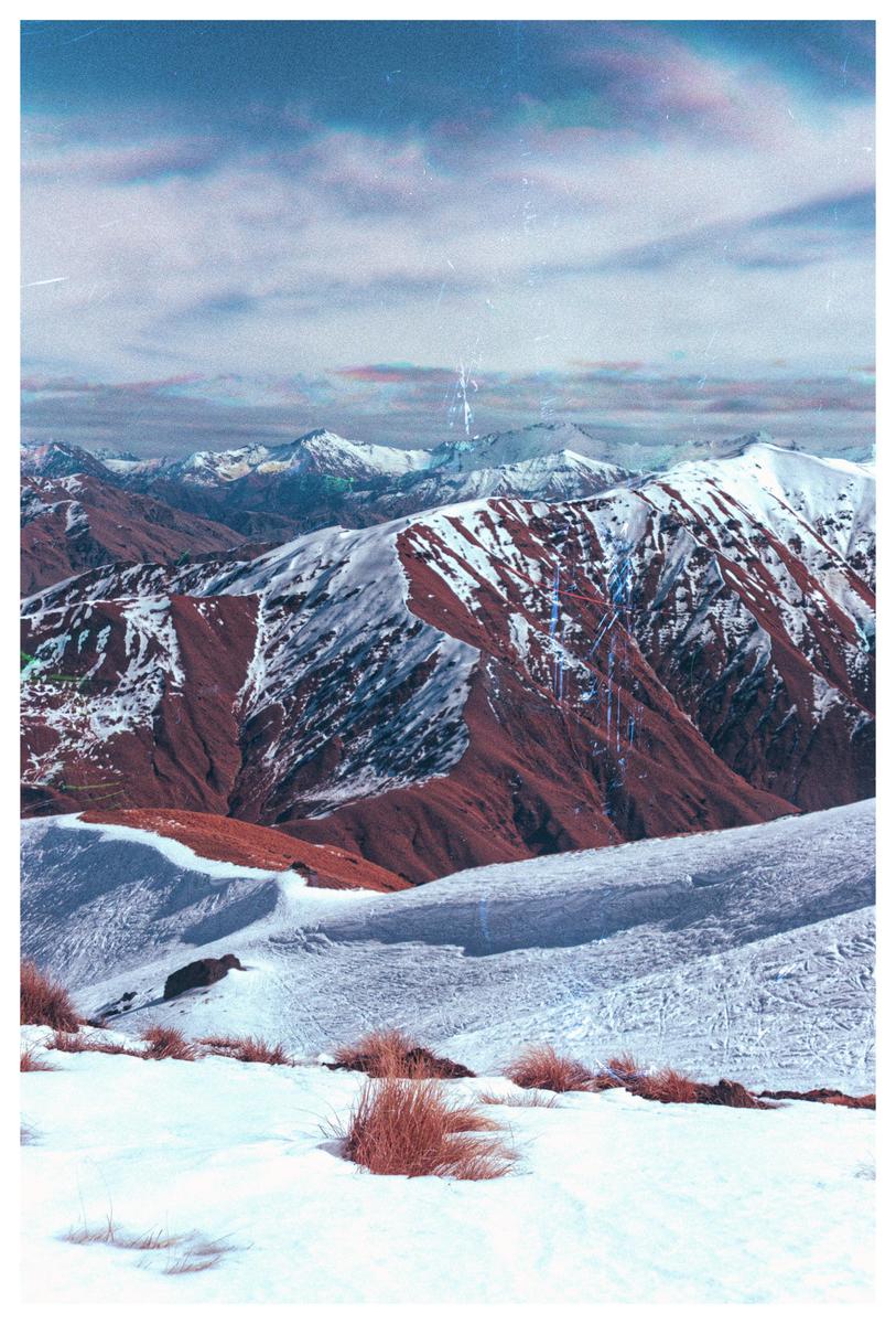 Snow-covered mountains with patches of red earth are visible under a cloudy sky.