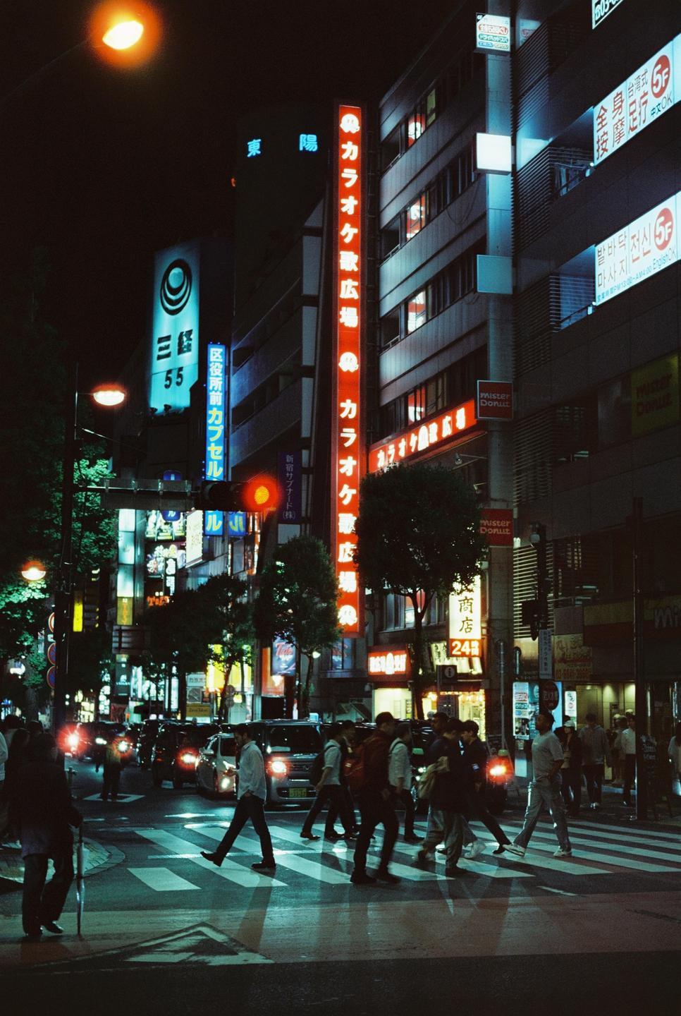 People cross a street at night in a city with tall buildings and neon signs.