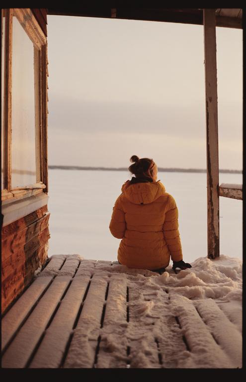 A woman in a yellow jacket sits on a snowy wooden deck, looking out at a frozen lake under a pale sky.