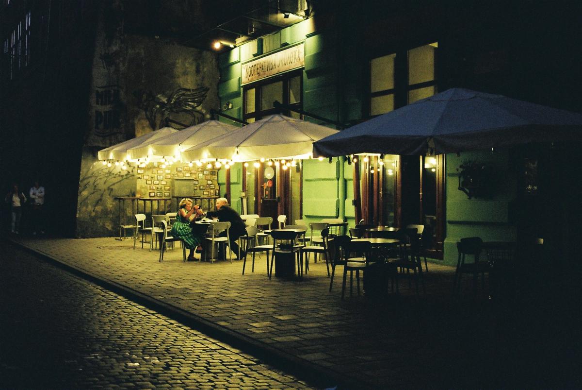 A couple sits at a table outside a restaurant at night, illuminated by string lights under an awning.