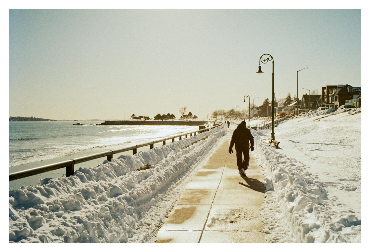 A person walks along a snow-lined path next to a body of water with houses on a hill in the background.