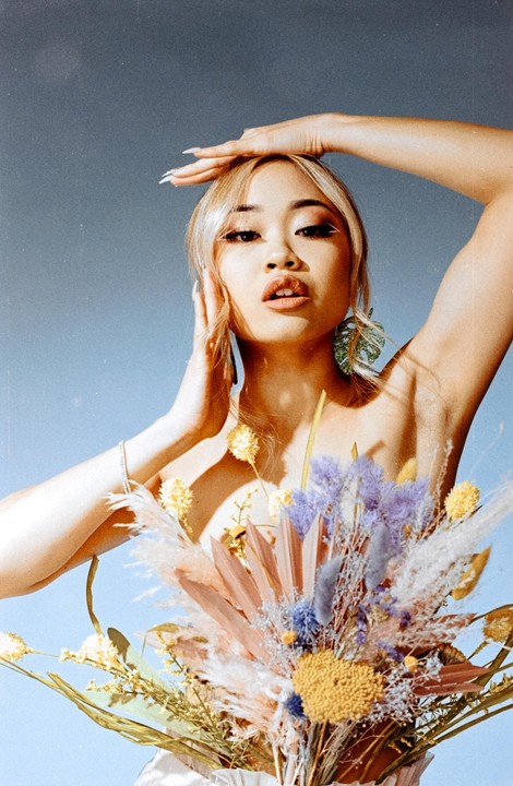 A woman with blonde hair and white eyeliner poses with a bouquet of dried flowers in front of a blue background.