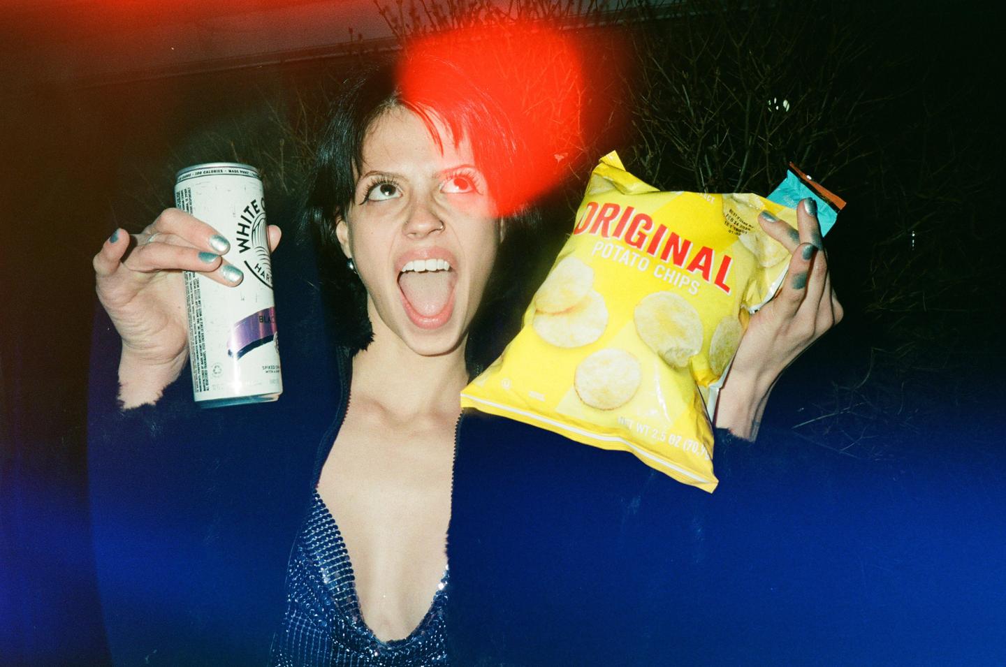 A woman with an open mouth holds a can and a bag of potato chips at night.