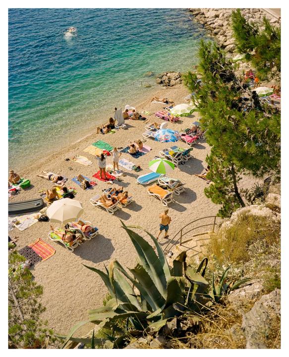 A high-angle view shows a crowded pebble beach with people sunbathing and swimming in the clear blue water.