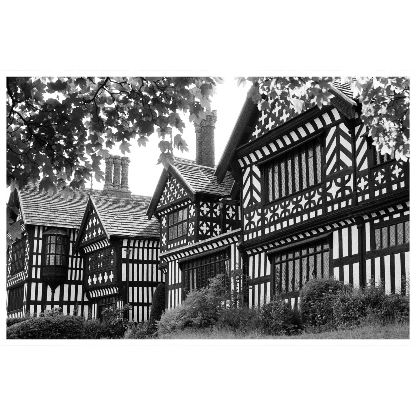 A black and white photograph shows the exterior of Bramall Hall, a historic timber-framed manor house, with trees framing the top of the image.