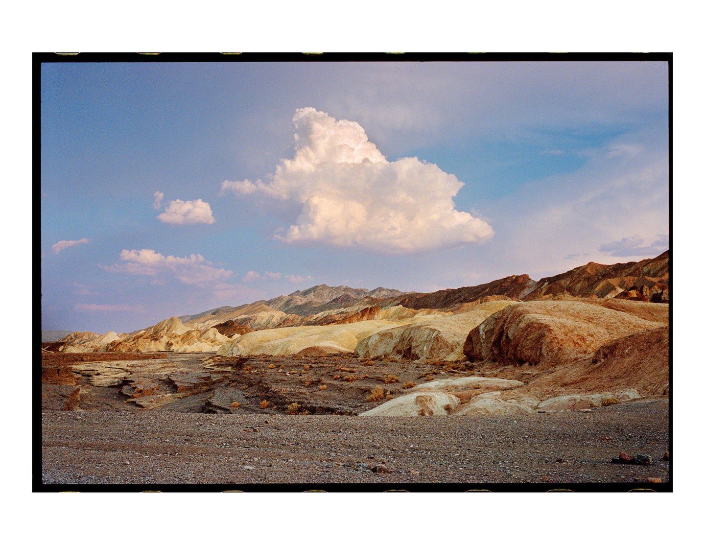 A vast desert landscape stretches under a blue sky with scattered clouds, featuring rugged mountains and dry, cracked earth.