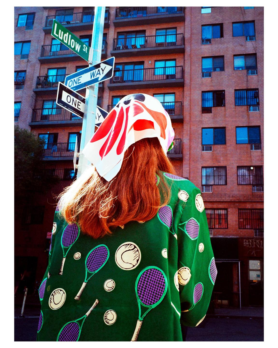 A woman with red hair and a green tennis-themed shirt stands on a city street with a building and street signs in the background.