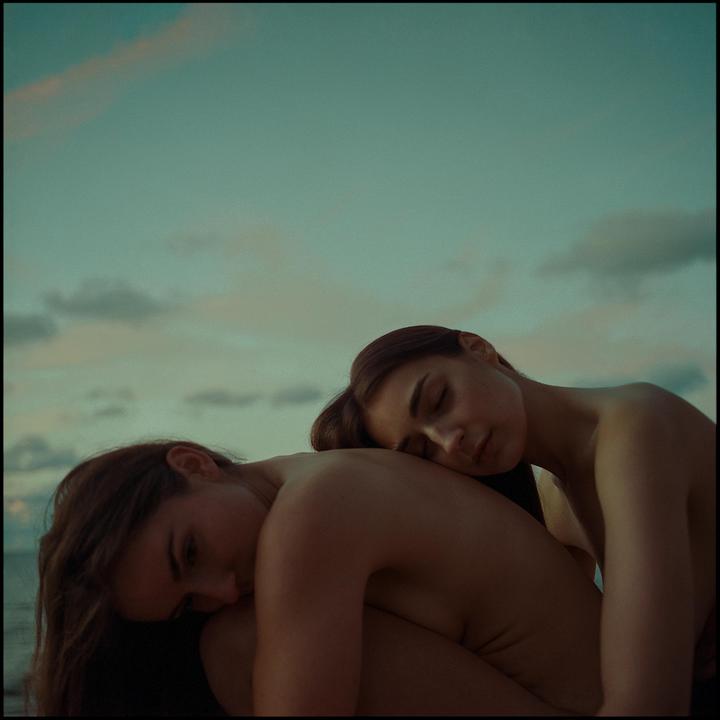 Two women are embracing each other with their eyes closed under a cloudy sky.