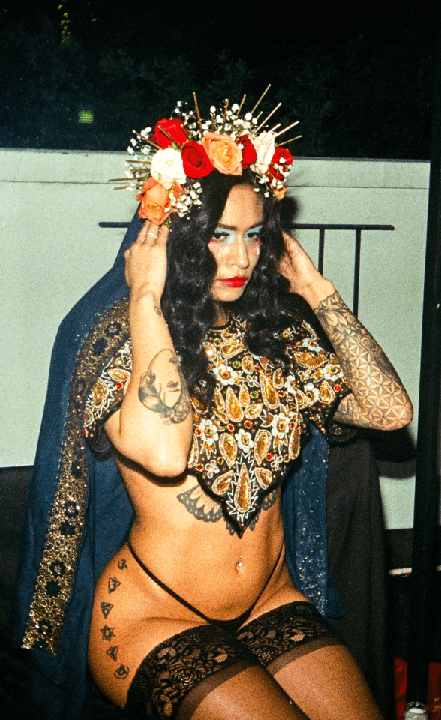 A woman with a flower crown, elaborate makeup, and tattoos poses for a portrait.