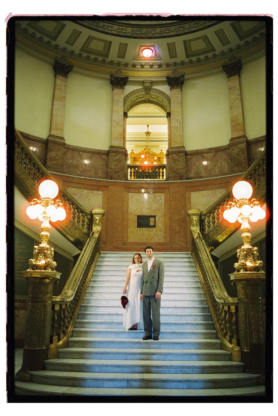 A couple stands on a grand marble staircase inside a building with ornate architecture and glowing light fixtures.