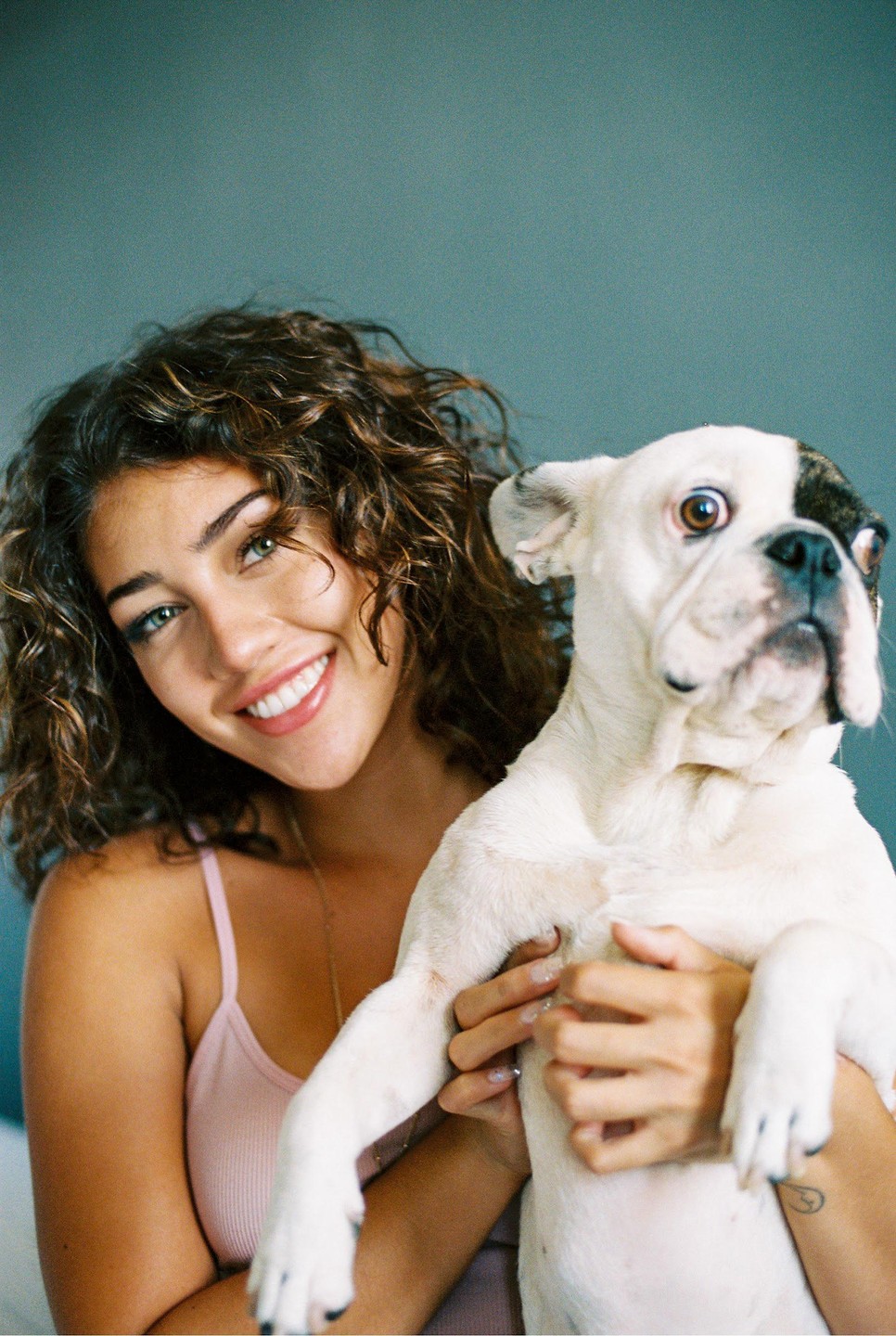 A woman with curly hair smiles while holding a white bulldog with a black patch over one eye.