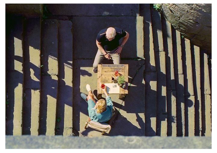 A man and a woman sit at a small table on stone steps with strong shadows.