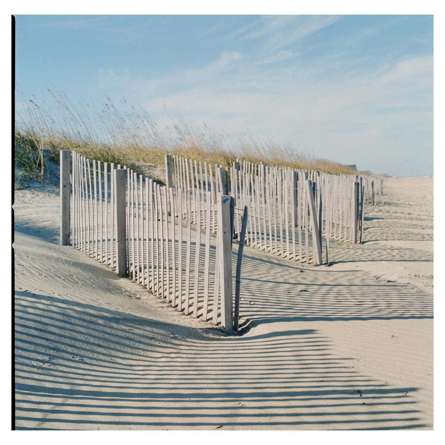 A wooden fence casts long shadows on a sandy beach with dune grass under a blue sky.