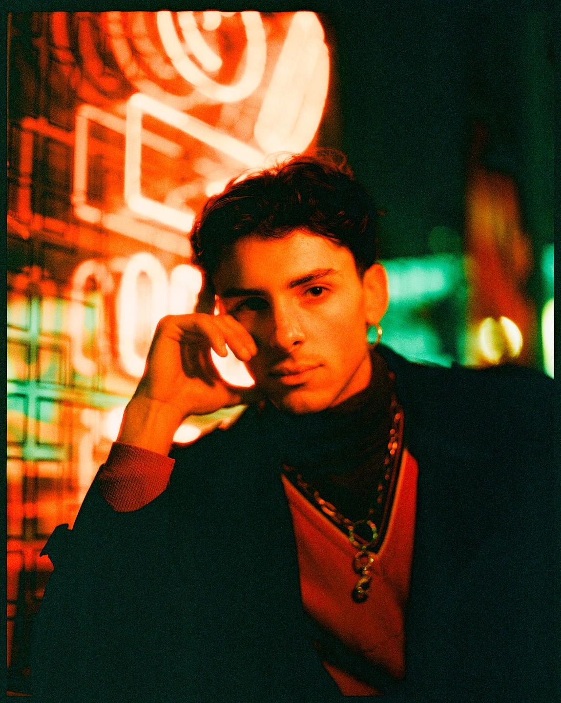 A man with dark hair and a gold earring looks at the viewer, illuminated by neon light.