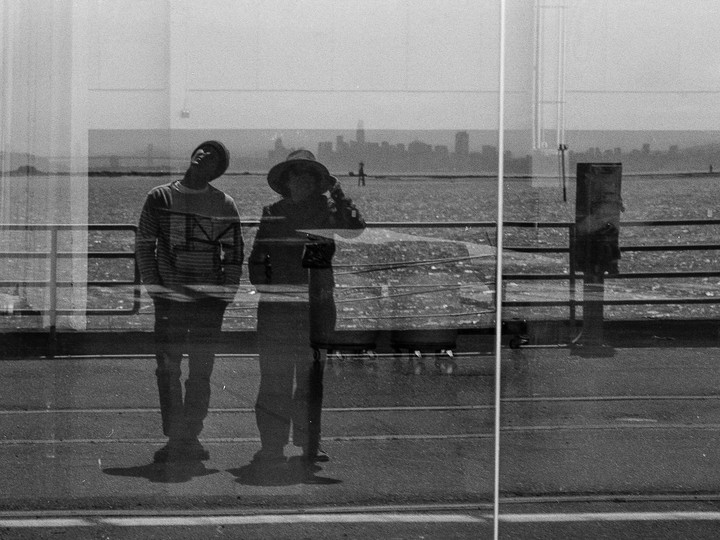 Two people stand by a railing overlooking a body of water with a city skyline in the distance, captured with a double exposure.