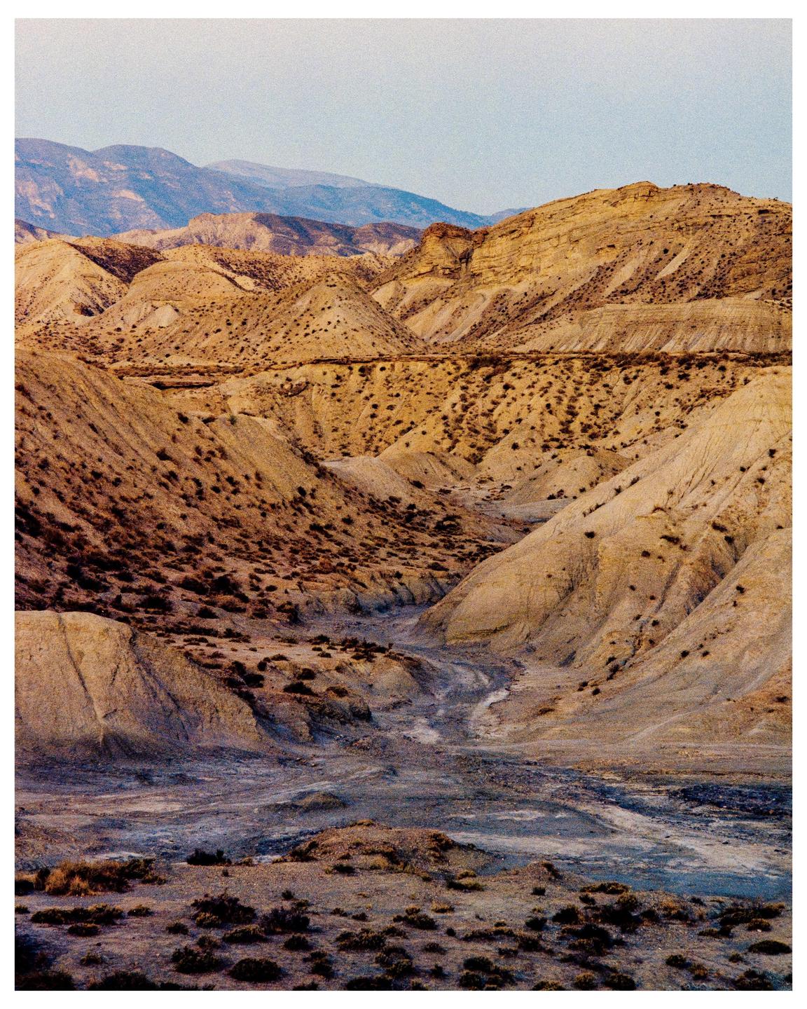 A dry riverbed winds through a desert landscape with rolling hills and mountains in the distance.