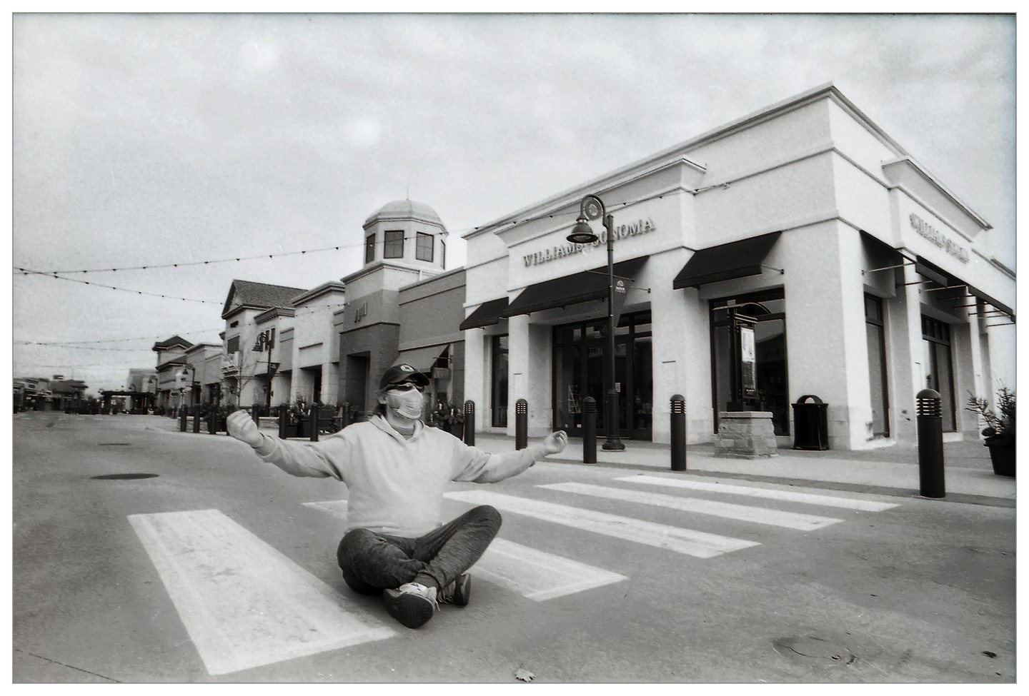 A man wearing a face mask sits cross-legged in the middle of a crosswalk in front of a row of stores.