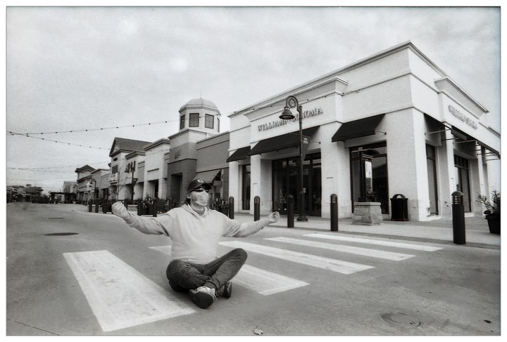 A man wearing a face mask sits cross-legged in the middle of a crosswalk in front of a row of stores.