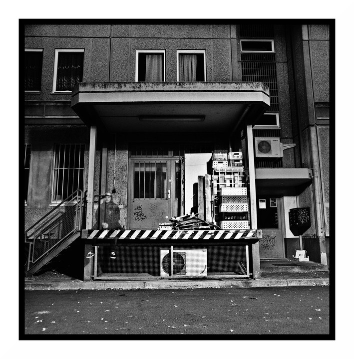 A black and white street photograph shows the entrance to a building with a loading dock, an air conditioning unit, and graffiti on the door.