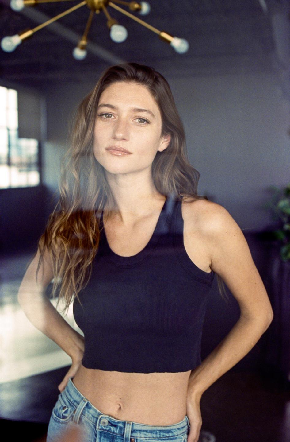 A woman with long hair and a dark tank top stands with her hands on her hips, looking directly at the viewer.