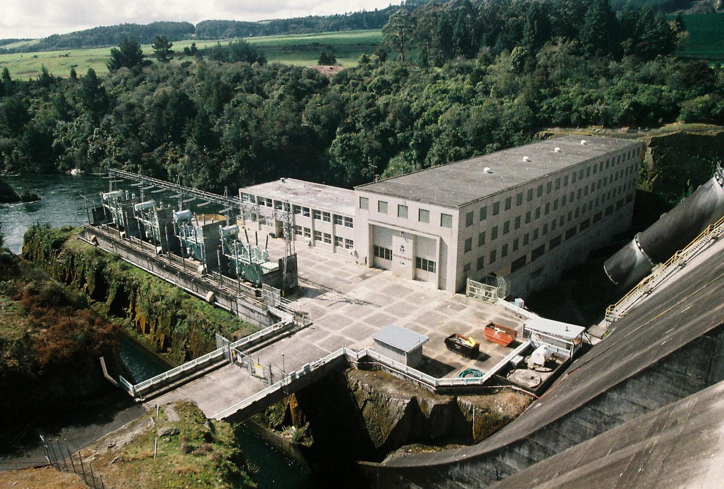 An aerial view shows the Whakamaru Dam and power station nestled among lush green hills and a river.
