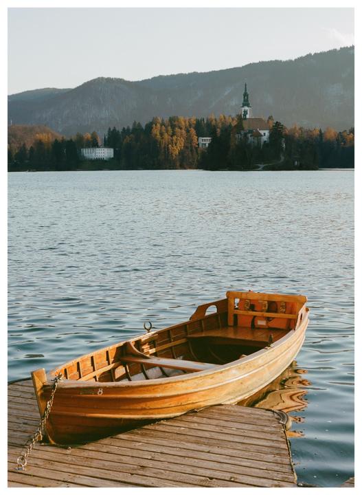 A wooden rowboat is tied to a dock on a lake with a church and mountains in the background.