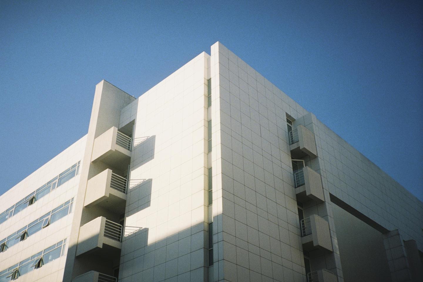 A modern building with balconies and many windows is photographed from below against a clear blue sky.