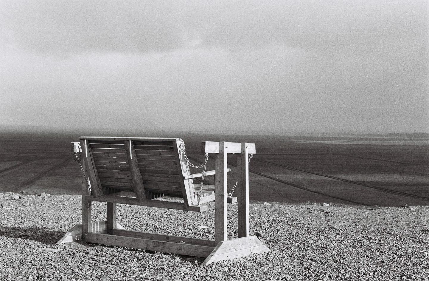 A wooden swing bench sits on a gravelly hill overlooking a vast, flat landscape under a cloudy sky.