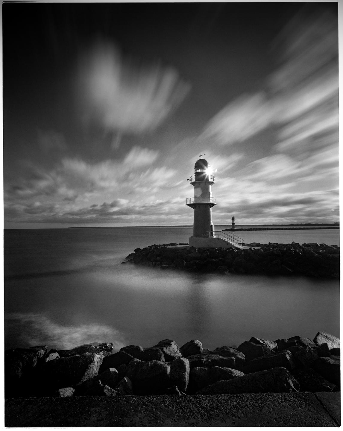 A lighthouse stands on a rocky jetty with blurred clouds in the sky and smooth water below.
