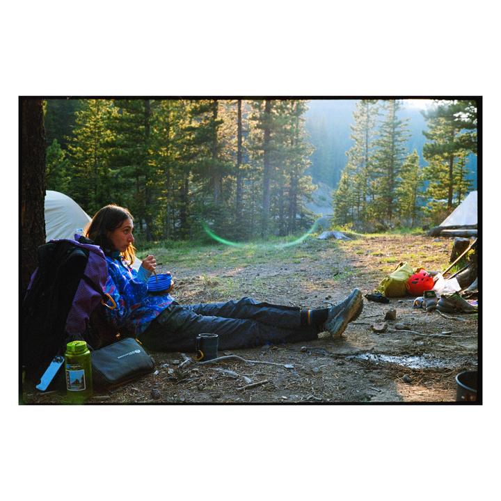 A woman sits on the ground next to a backpack and tent, eating from a bowl in a forest.