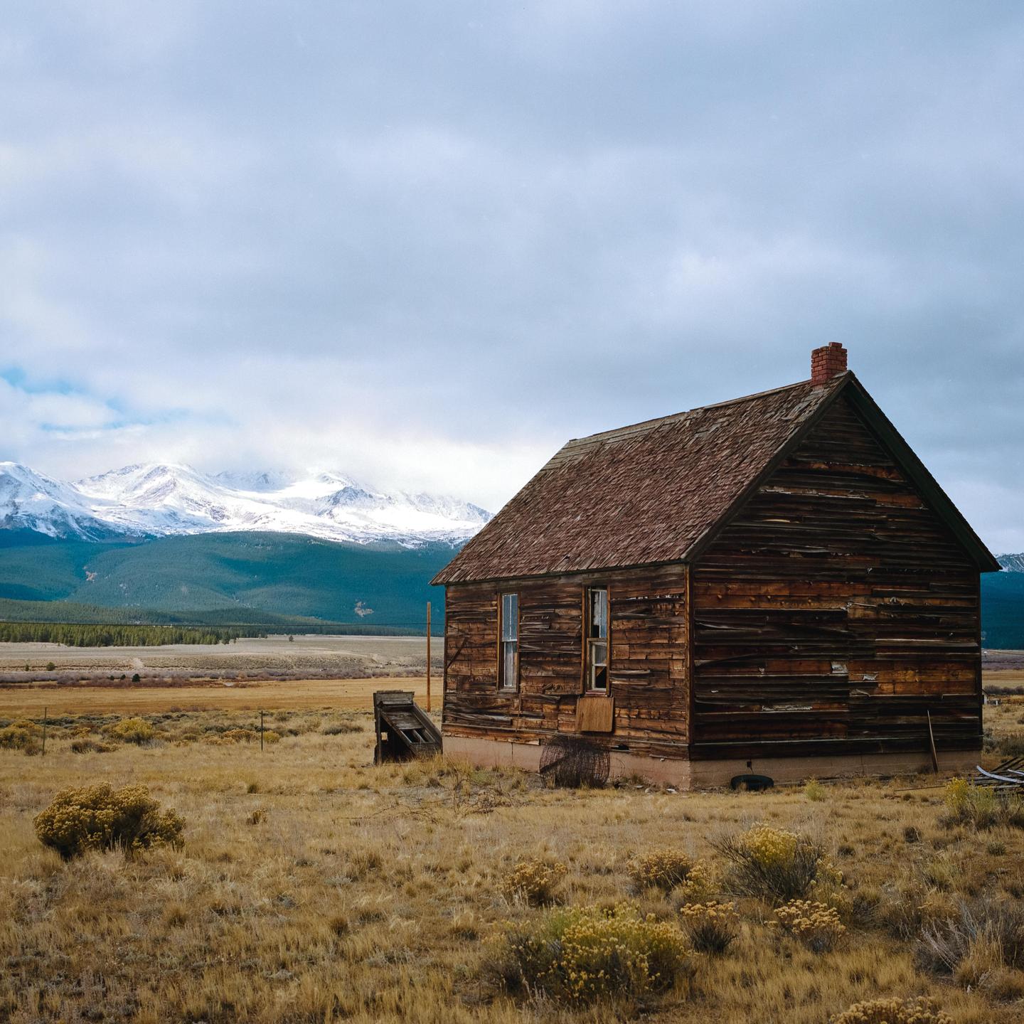 An old wooden house stands in a field with snow-capped mountains in the background under a cloudy sky.