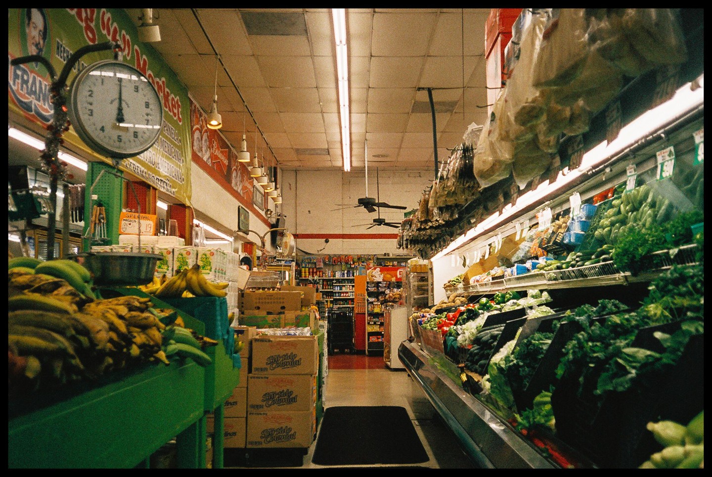 A wide shot of a supermarket aisle shows produce on the right and bananas on the left, with a clock hanging above the bananas.