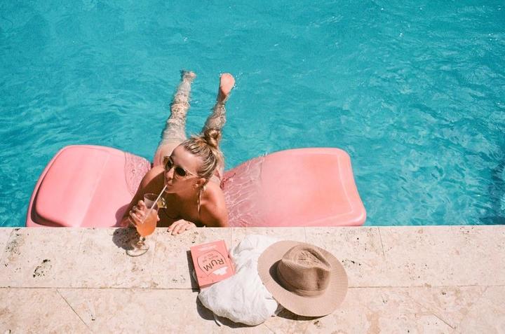 A woman floats on a pink raft in a swimming pool, drinking from a glass with a straw, with a book and hat on the pool deck.
