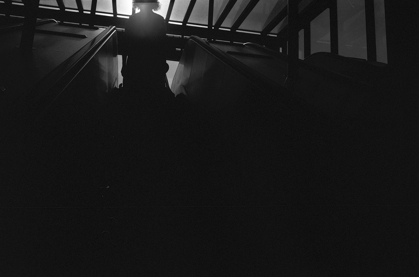 A person stands at the top of an escalator, silhouetted against a bright window.