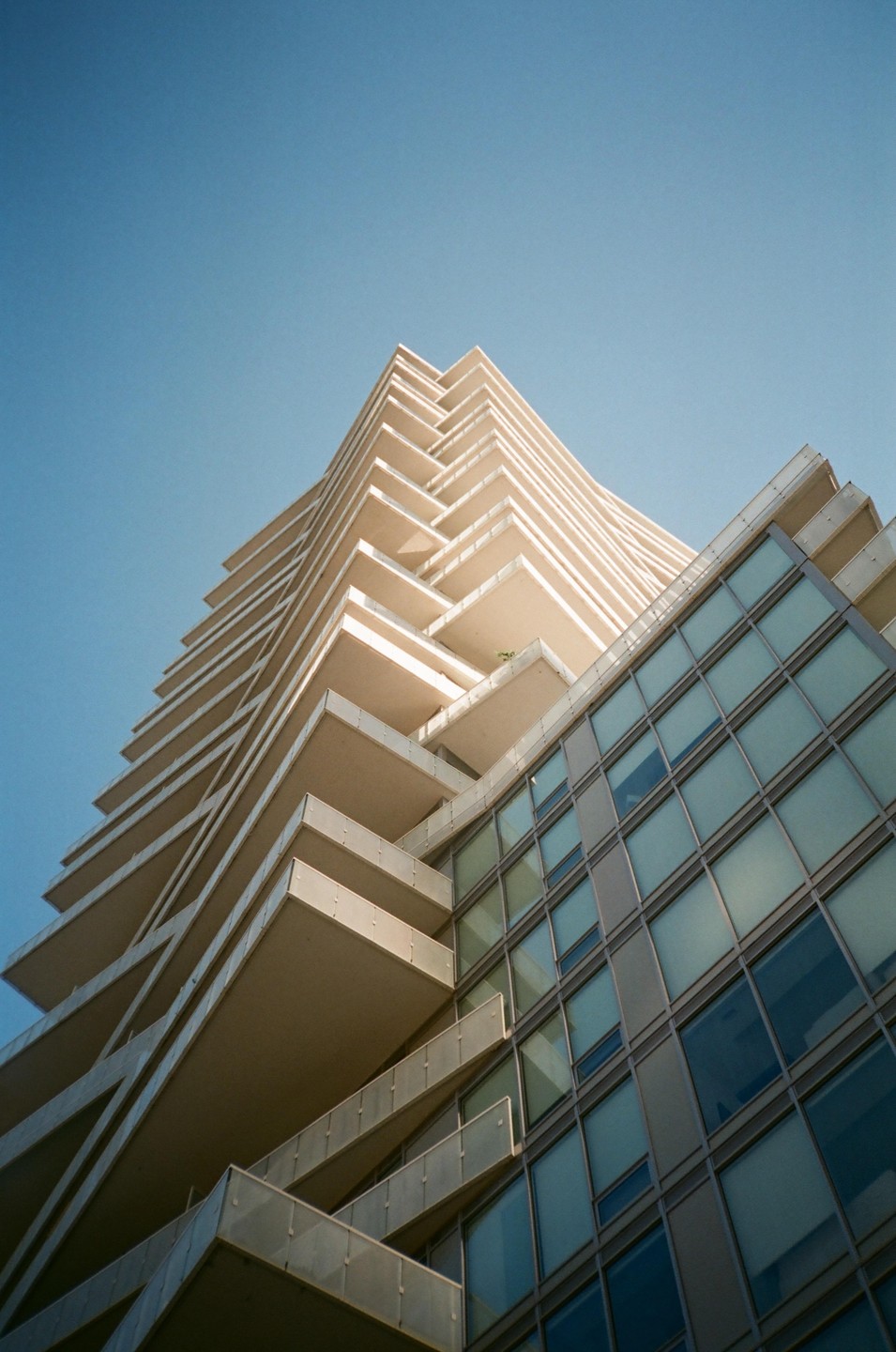A modern high-rise building with balconies and a glass facade stands against a clear blue sky.
