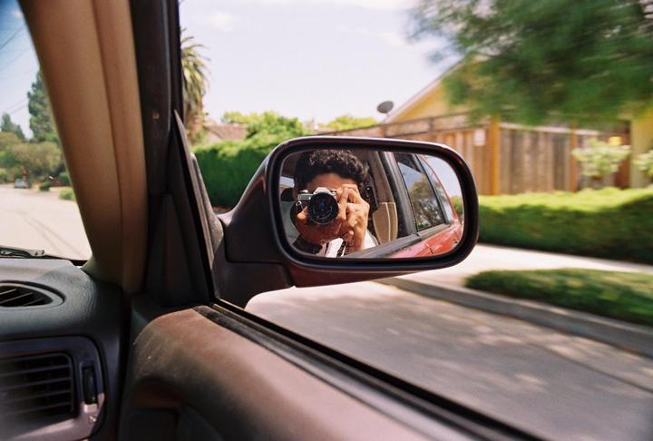 A person is visible in the side mirror of a car, holding a camera and taking a self-portrait.