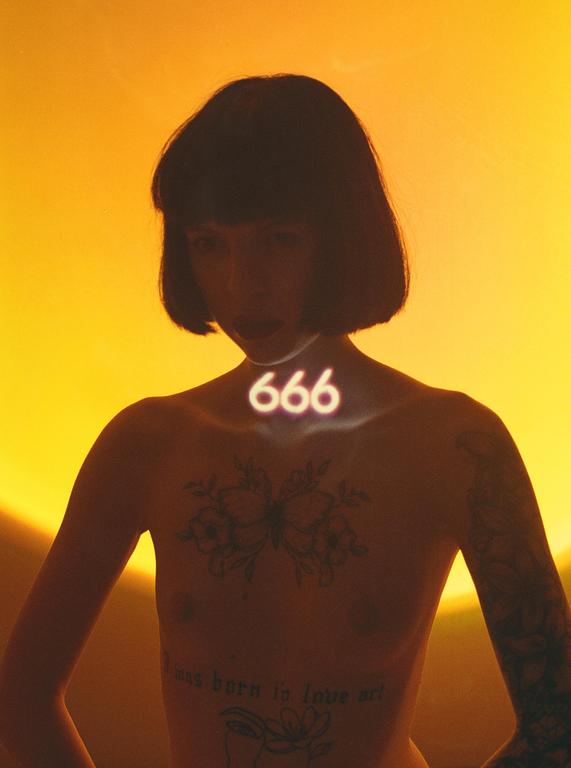 A woman with short hair and tattoos on her chest and arms has the number 666 projected onto her neck.