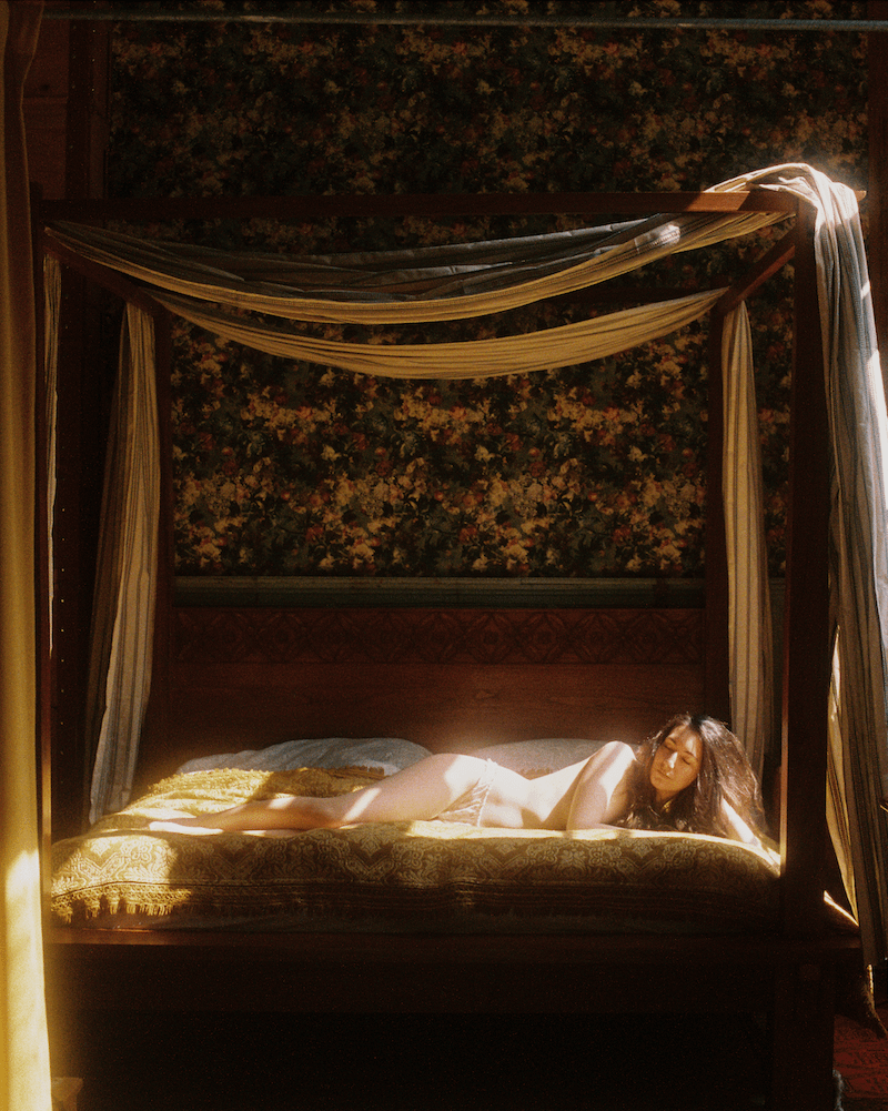 A woman lies on a four-poster bed with light shining on her body.