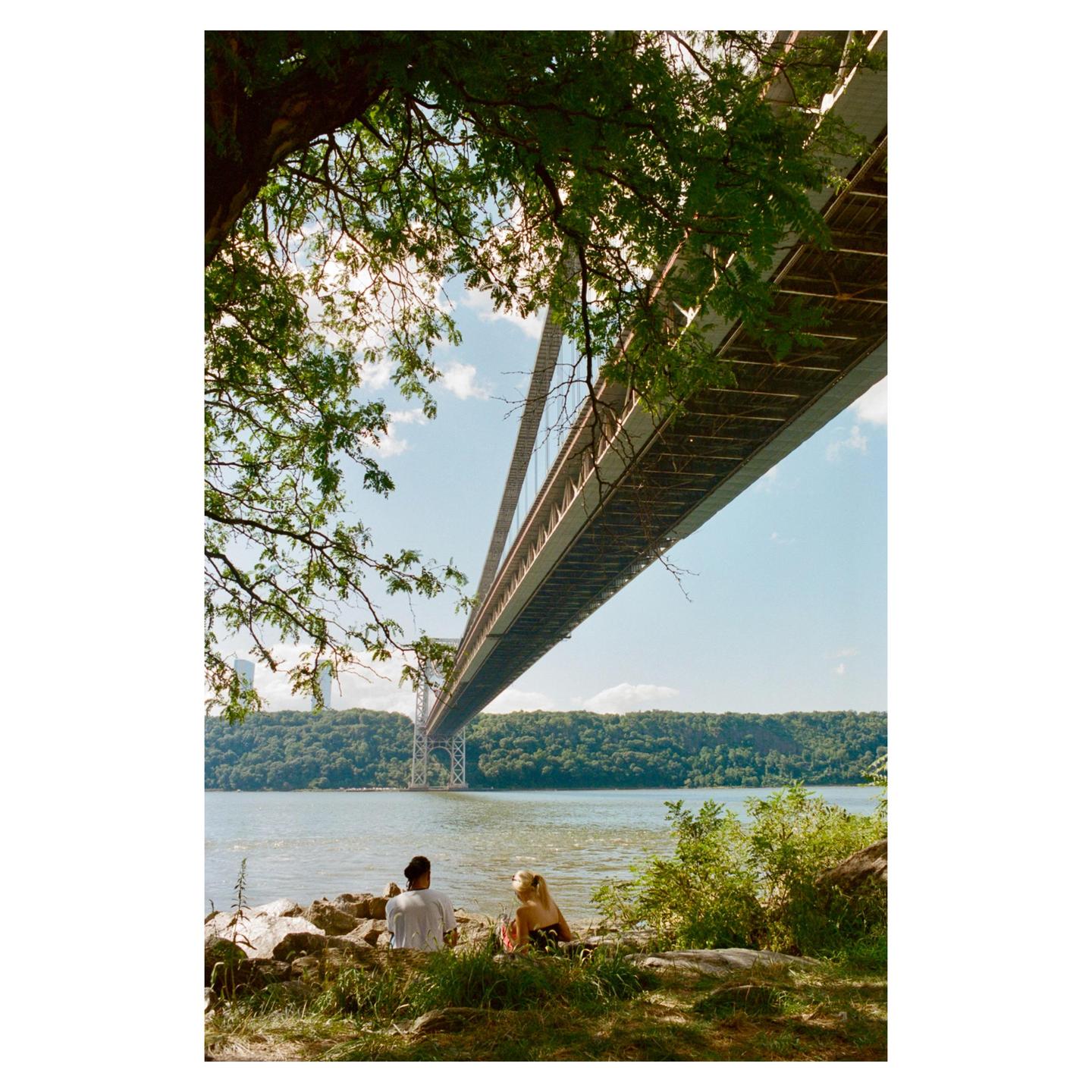A couple sits on rocks by a river under a large suspension bridge.