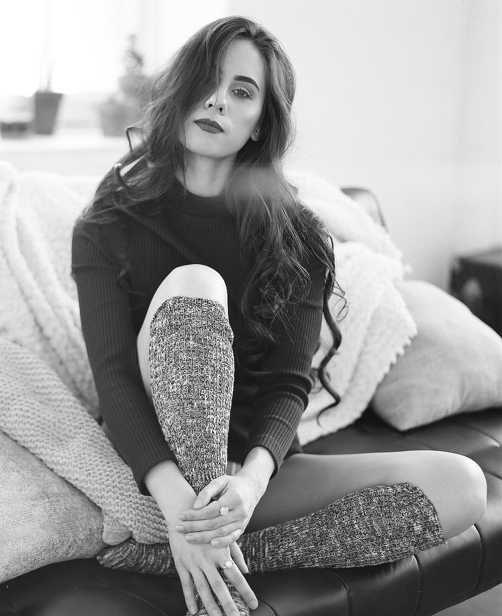 A woman with long hair and dark lipstick sits on a couch with her legs crossed, wearing a sweater and leg warmers.