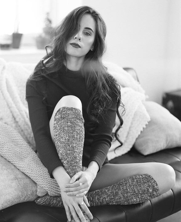 A woman with long hair and dark lipstick sits on a couch with her legs crossed, wearing a sweater and leg warmers.