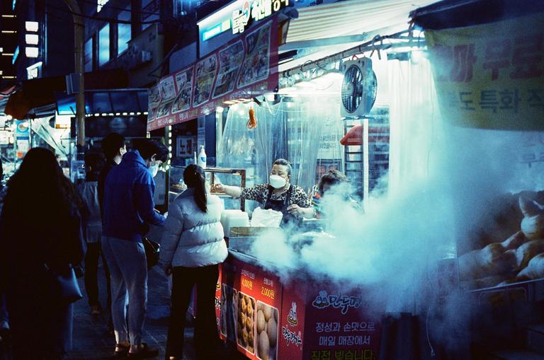 A woman wearing a face mask serves food from a steaming street stall at night while people wait in line.