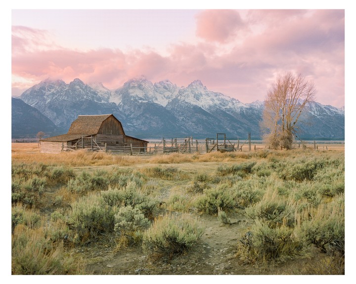 A wooden barn stands in a field with snow-capped mountains and a pink sky in the background.