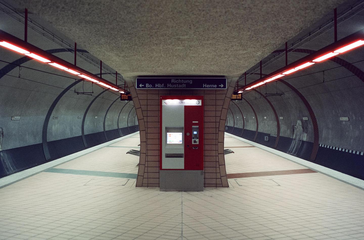 A subway platform with a ticket machine in the center is illuminated by red fluorescent lights.