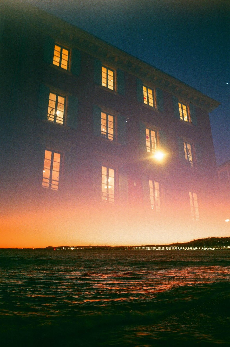 A double exposure shows a building with illuminated windows superimposed over a body of water at sunset.