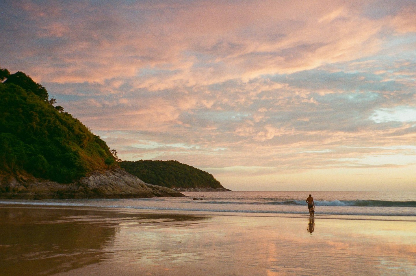 A man and a boy stand in the shallow water of a beach at sunset, with a lush green hill on the left.