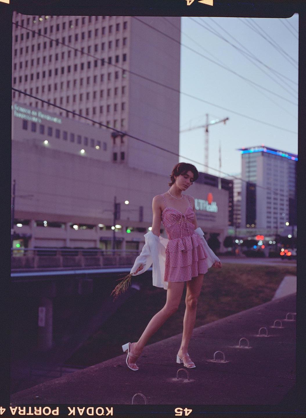 A woman in a pink gingham dress and white shirt stands on a ledge with city buildings in the background.