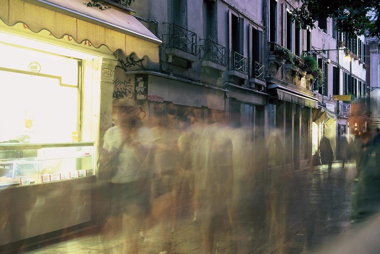 A long exposure shot captures blurred figures walking past a brightly lit storefront and buildings at night.
