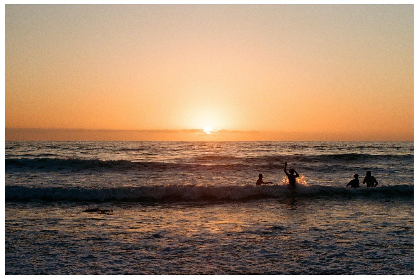 Four people are silhouetted in the ocean as the sun sets over the water.
